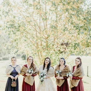 May include: Five women in a line, wearing burgundy and navy blue dresses with faux fur wraps. The woman in the middle is wearing a white lace dress and holding a bouquet of white flowers. They are standing in front of a tree with yellow leaves.