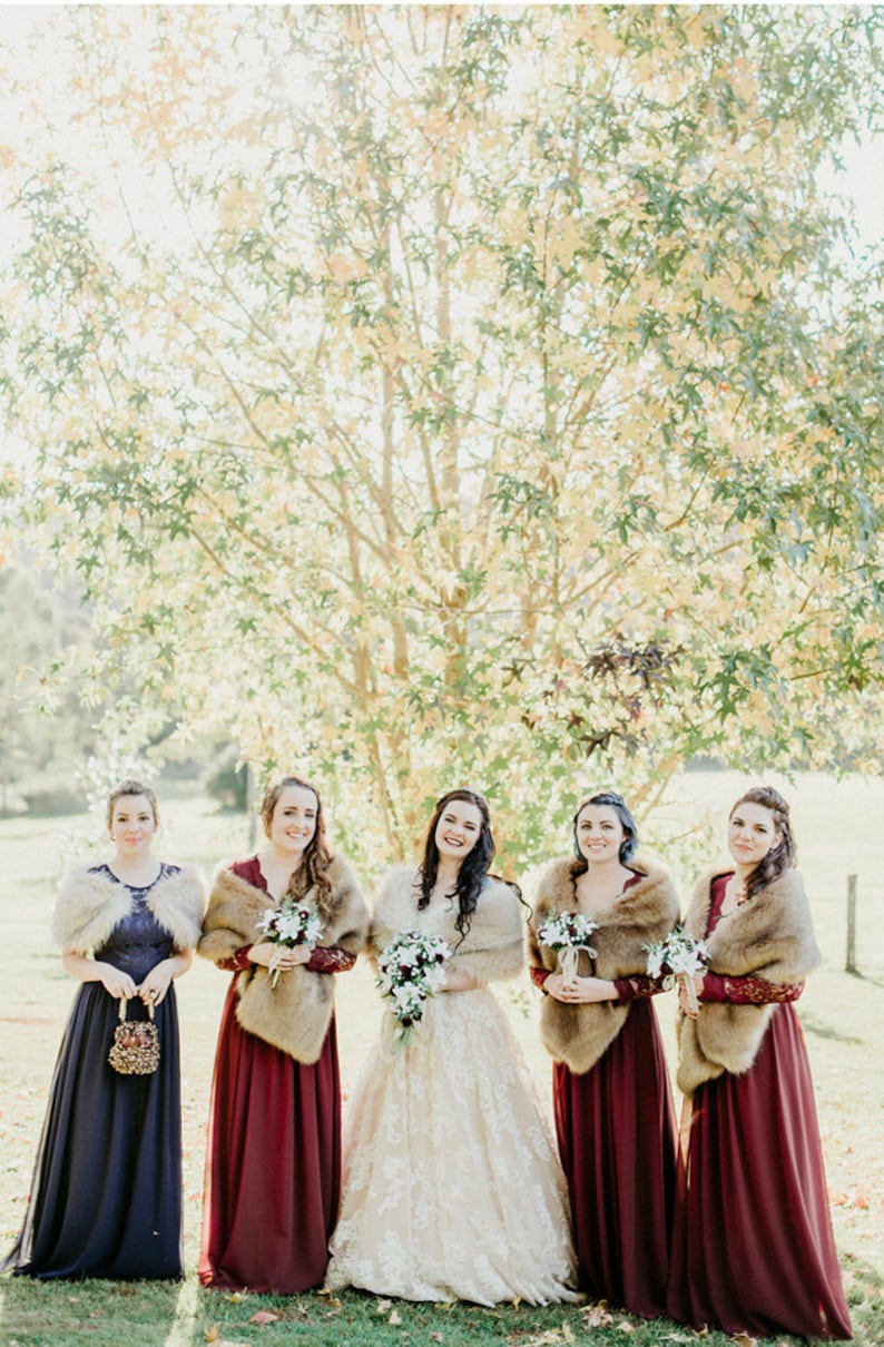 May include: Five women in a line, wearing burgundy and navy blue dresses with faux fur wraps. The woman in the middle is wearing a white lace dress and holding a bouquet of white flowers. They are standing in front of a tree with yellow leaves.