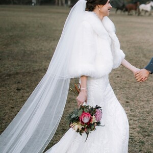 May include: A bride wearing a white lace wedding dress with a white faux fur shrug and a long white veil. She is holding a bouquet of pink and white flowers.