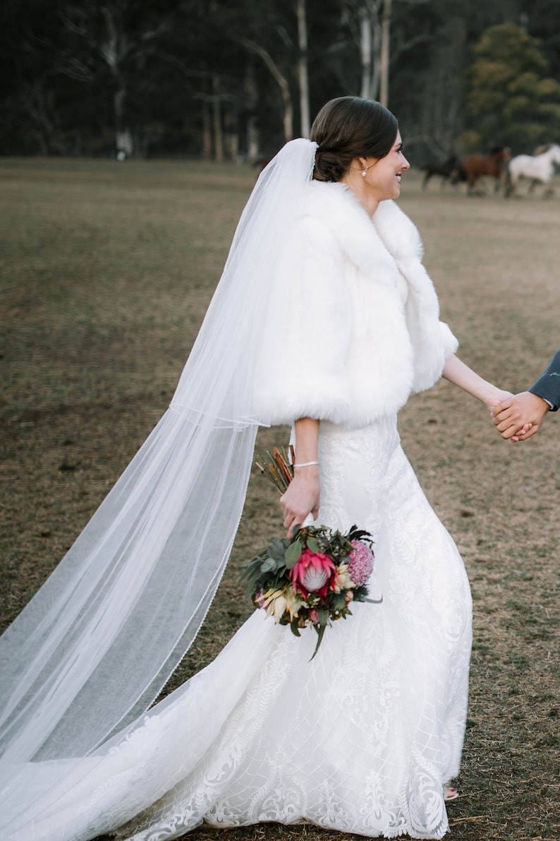 May include: A bride wearing a white lace wedding dress with a white faux fur shrug and a long white veil. She is holding a bouquet of pink and white flowers.
