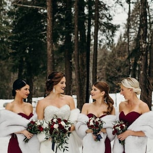 May include: Four bridesmaids in burgundy dresses with white faux fur wraps stand in a snowy forest. The bride is in the center wearing a white strapless wedding dress with a white faux fur wrap and a bouquet of red and white flowers.