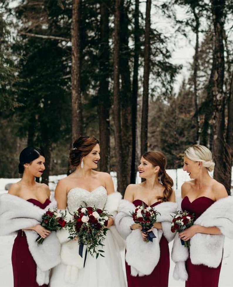 May include: Four bridesmaids in burgundy dresses with white faux fur wraps stand in a snowy forest. The bride is in the center wearing a white strapless wedding dress with a white faux fur wrap and a bouquet of red and white flowers.