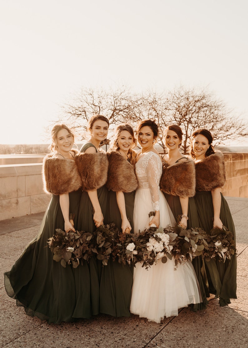 May include: Seven women in green dresses and faux fur wraps stand together holding bouquets of greenery and white flowers. The woman in the center is wearing a white lace wedding dress.