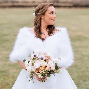 May include: A bride in a white wedding dress with a white fur shawl holds a bouquet of white, pink, and peach flowers.