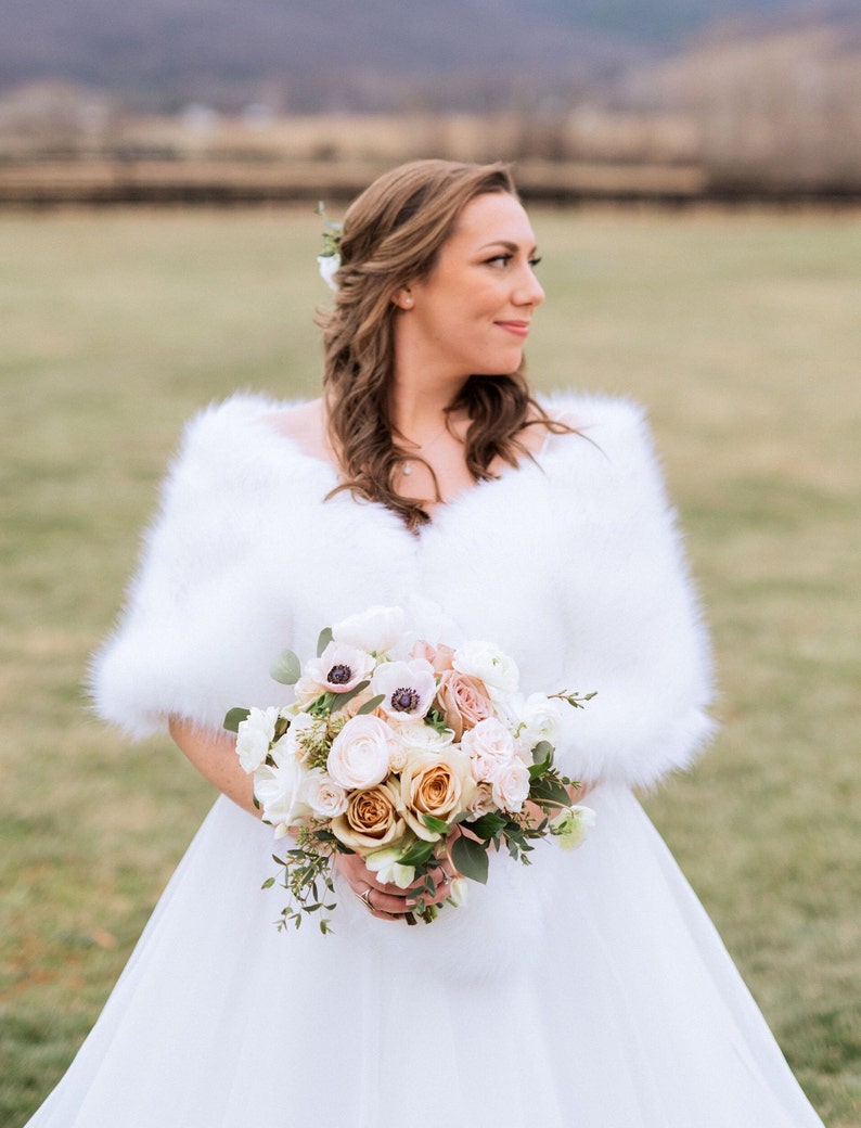 May include: A bride in a white wedding dress with a white fur shawl holds a bouquet of white, pink, and peach flowers.