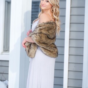 May include: A woman wearing a long, light gray dress with a brown faux fur shawl draped over her shoulders. She is standing in front of a white and gray house with snow on the ground.