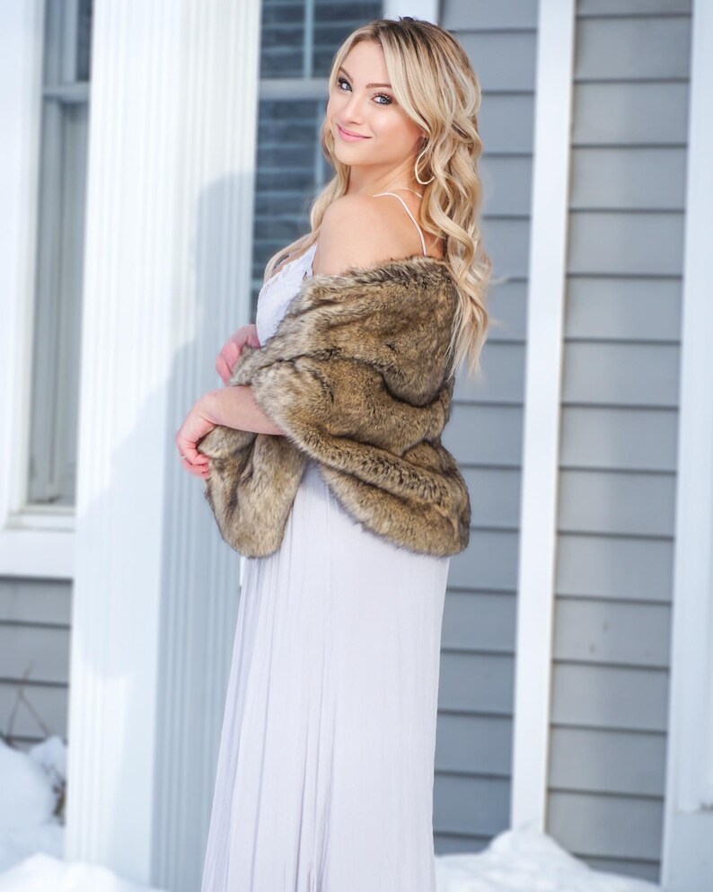 May include: A woman wearing a long, light gray dress with a brown faux fur shawl draped over her shoulders. She is standing in front of a white and gray house with snow on the ground.