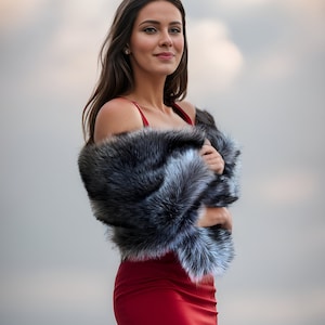 May include: A woman wearing a red dress with a black and white fur shawl. She is standing in front of a cloudy sky.