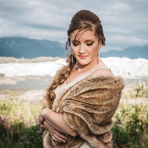 May include: A bride wearing a white wedding dress with a brown fur shawl. She is standing in a field with mountains in the background.