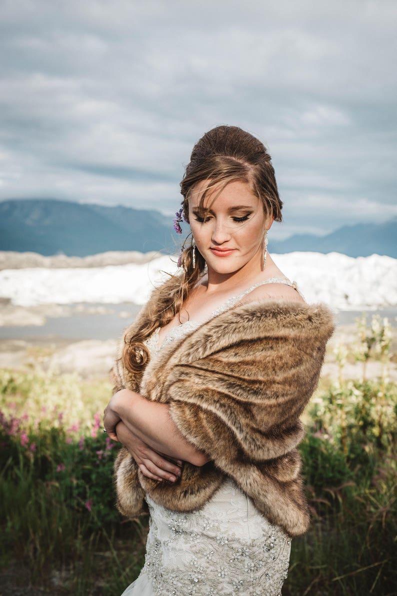May include: A bride wearing a white wedding dress with a brown fur shawl. She is standing in a field with mountains in the background.