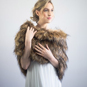 May include: A woman wearing a white dress with a brown and black faux fur shawl. The shawl is draped around her shoulders and she is holding it in front of her.