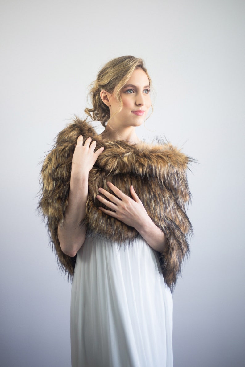 May include: A woman wearing a white dress with a brown and black faux fur shawl. The shawl is draped around her shoulders and she is holding it in front of her.
