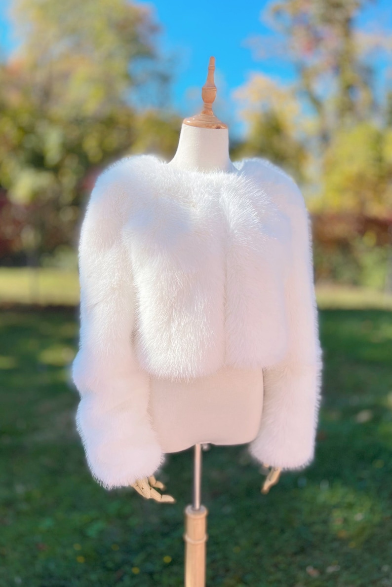 May include: A white faux fur cropped jacket with long sleeves. The jacket is displayed on a mannequin against a blurred background of green grass and trees.