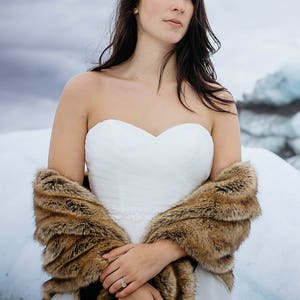 May include: A bride wearing a white strapless wedding dress with a brown fur shawl. She is standing in a snowy landscape.