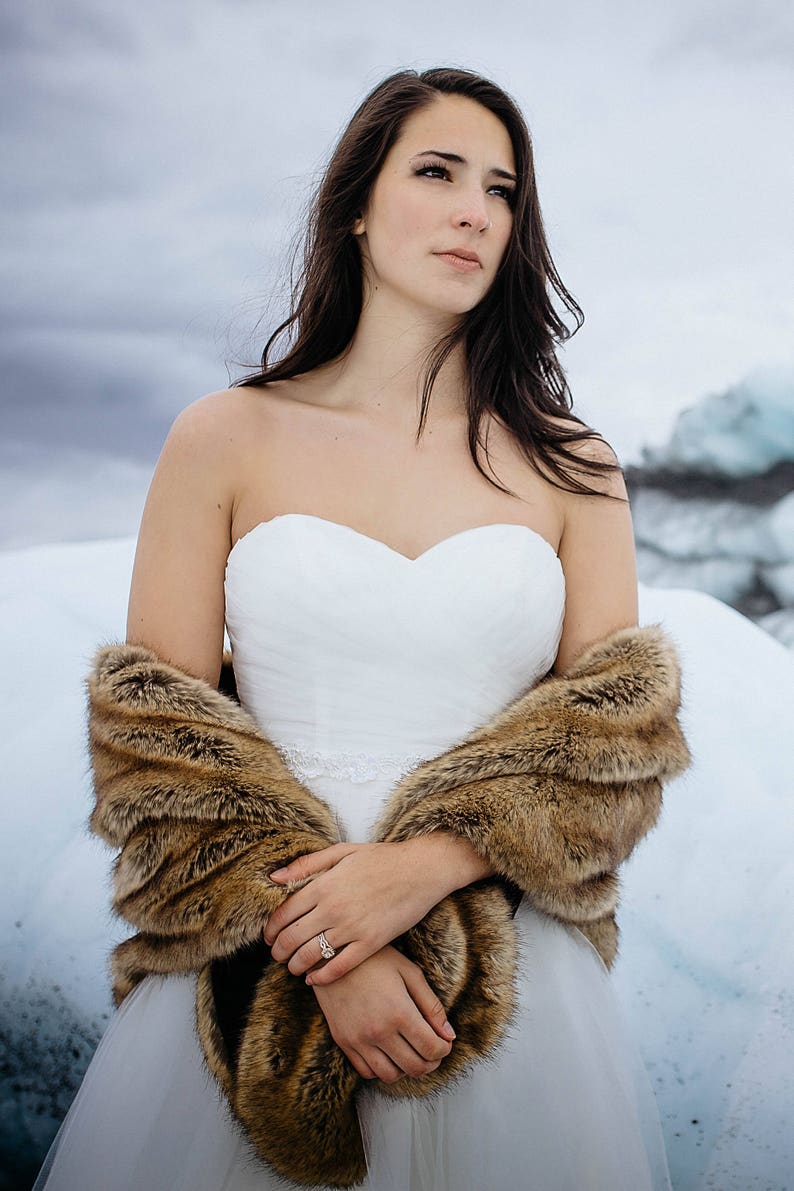 May include: A bride wearing a white strapless wedding dress with a brown fur shawl. She is standing in a snowy landscape.