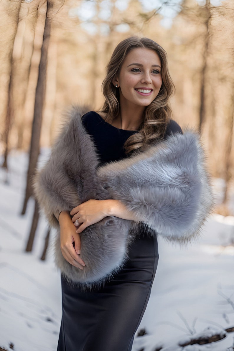May include: A woman wearing a gray faux fur shawl over a black dress. She is standing in a snowy forest with trees in the background.