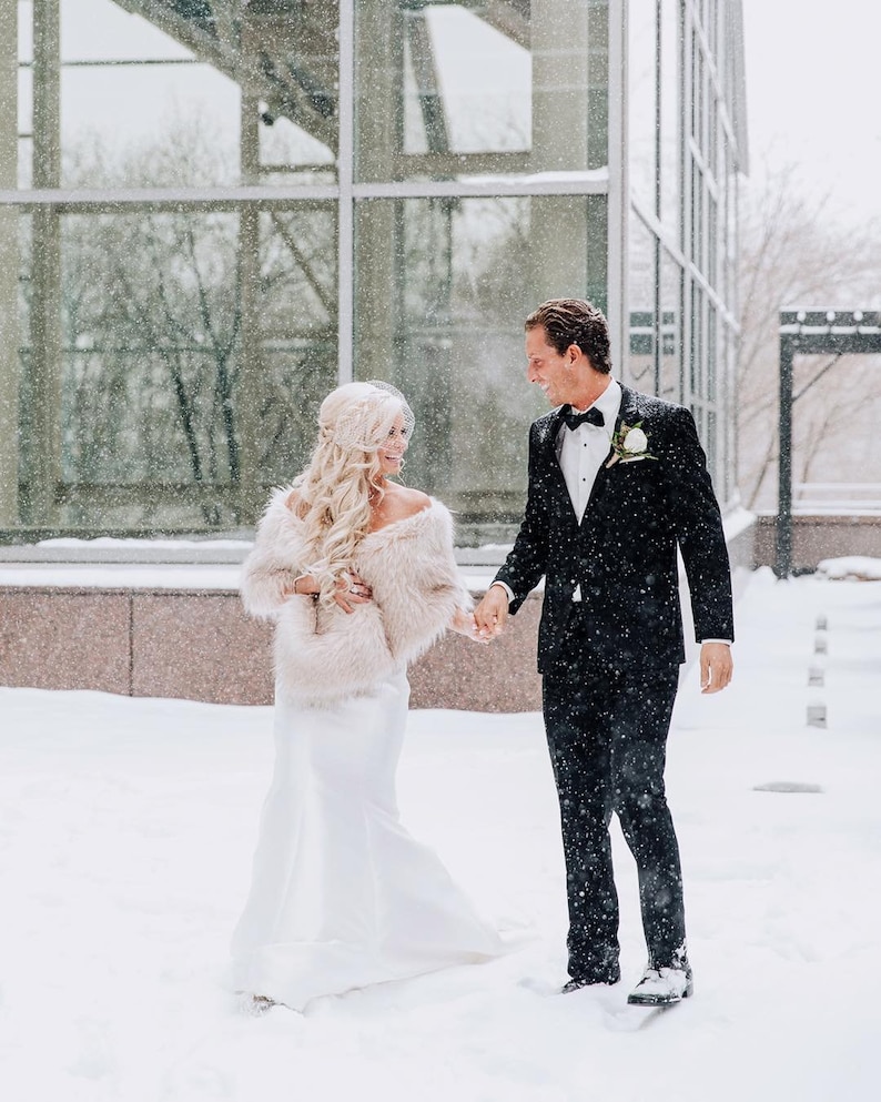 May include: A bride and groom walking hand-in-hand in the snow. The bride is wearing a white wedding dress with a fur shawl and the groom is wearing a black tuxedo. The couple is smiling and looking at each other.
