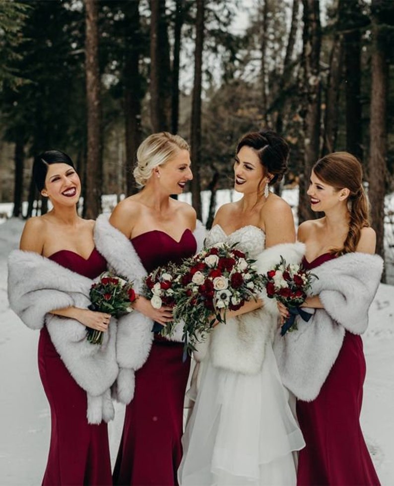 May include: Four bridesmaids in burgundy dresses with white faux fur wraps stand in a snowy forest. The bride in a white dress with a faux fur wrap holds a bouquet of red and white flowers.