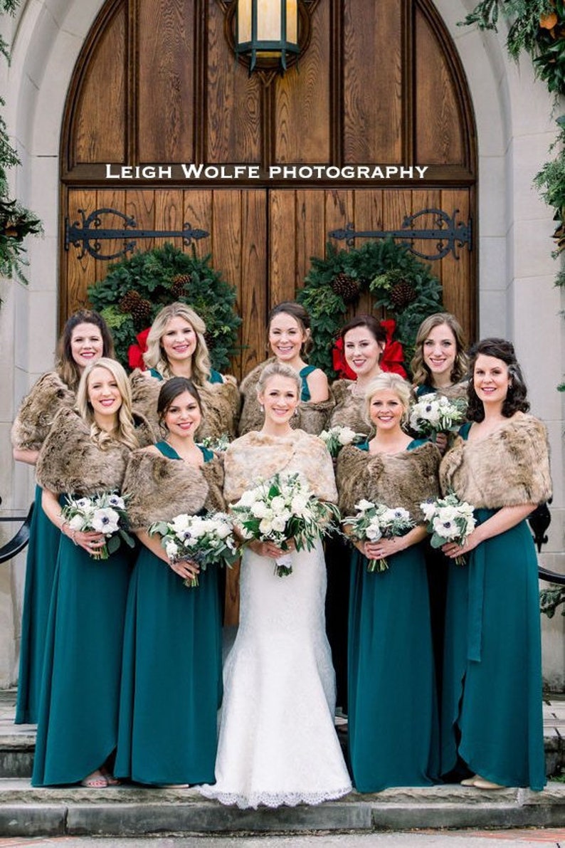 May include: A group of bridesmaids wearing teal green dresses and brown faux fur wraps stand in front of a wooden door with wreaths. The bride is in the center wearing a white lace wedding dress and holding a bouquet of white flowers.