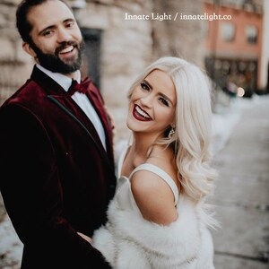 May include: A couple poses for a wedding photo. The groom is wearing a burgundy velvet suit jacket with a black bow tie. The bride is wearing a white dress with a white fur shawl. The photo is taken in an urban setting with a brick wall in the background.