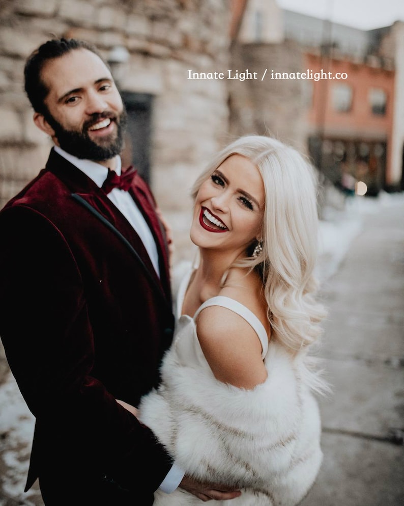 May include: A couple poses for a wedding photo. The groom is wearing a burgundy velvet suit jacket with a black bow tie. The bride is wearing a white dress with a white fur shawl. The photo is taken in an urban setting with a brick wall in the background.