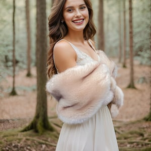 May include: A woman in a white dress with a light brown fur shawl standing in a forest. She is smiling and looking at the camera.