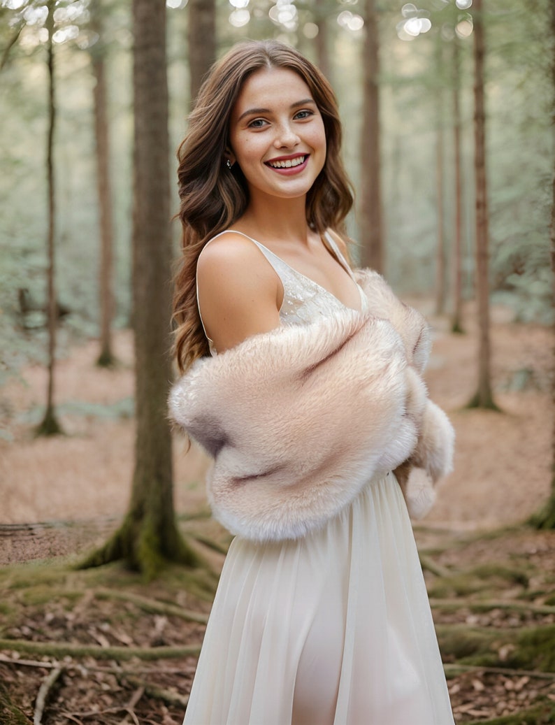 May include: A woman in a white dress with a light brown fur shawl standing in a forest. She is smiling and looking at the camera.