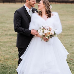 May include: A bride and groom pose for a wedding portrait outdoors. The bride is wearing a white wedding dress with a fur shawl and a bouquet of flowers. The groom is wearing a black suit and a green tie. They are standing in a grassy field with a fence in the background.