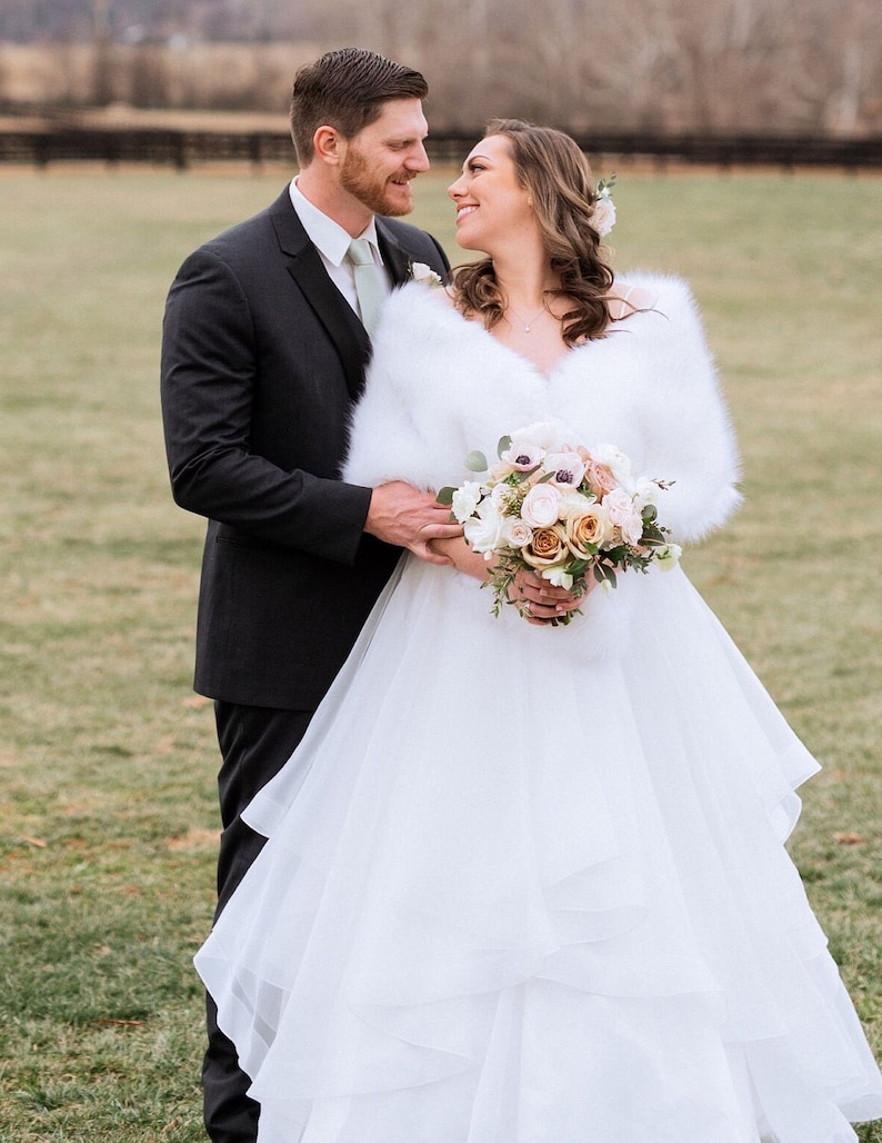 May include: A bride and groom pose for a wedding portrait outdoors. The bride is wearing a white wedding dress with a fur shawl and a bouquet of flowers. The groom is wearing a black suit and a green tie. They are standing in a grassy field with a fence in the background.