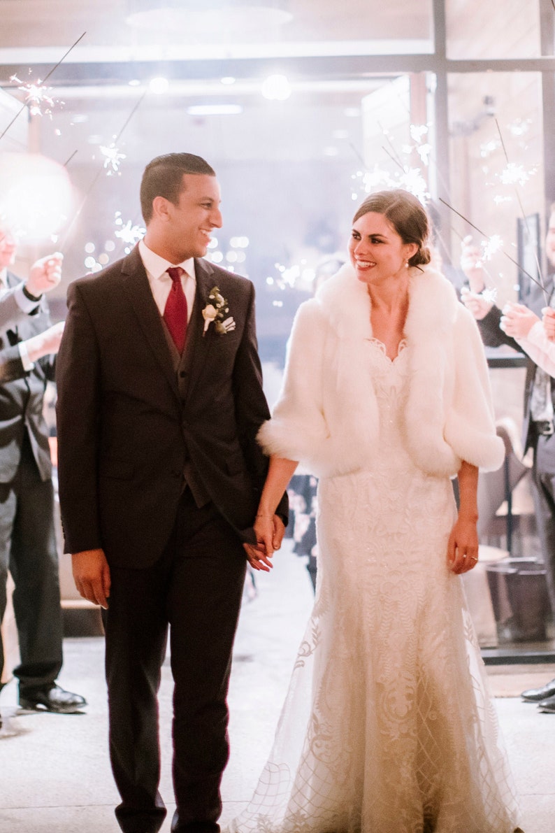 May include: A bride and groom walking out of a building, holding hands. The bride is wearing a white wedding dress with a white fur shawl. The groom is wearing a dark suit with a burgundy tie. They are surrounded by people holding sparklers.