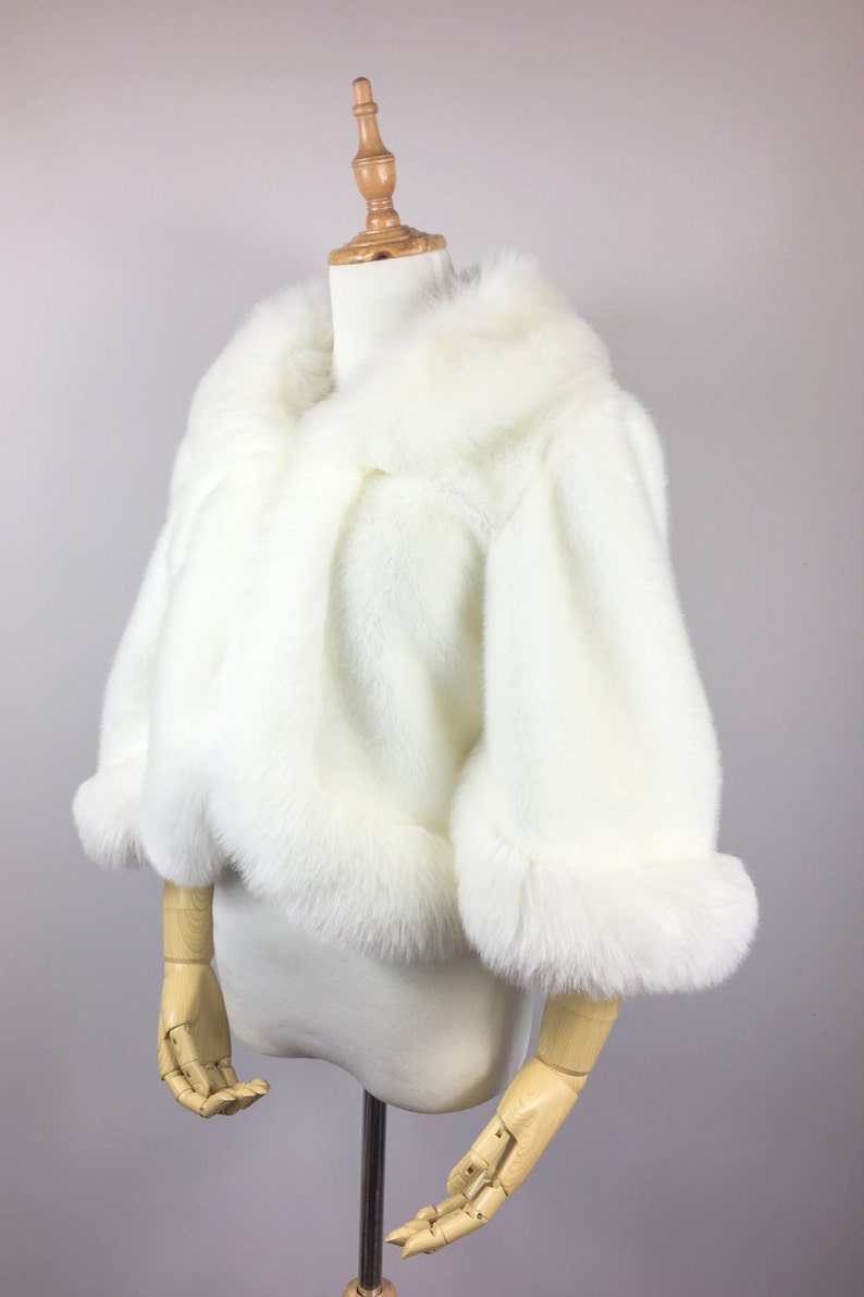 May include: A white faux fur shrug with a shawl collar and long sleeves. The shrug is made of soft, fluffy faux fur and has a relaxed fit.