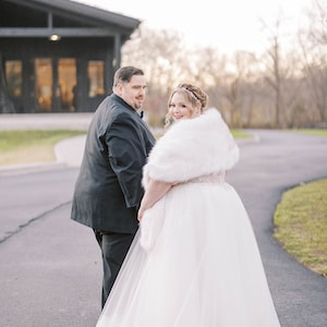 May include: A bride and groom walking down a paved path. The bride is wearing a white wedding dress with a fur shawl and a tiara. The groom is wearing a black suit. They are walking away from the camera and smiling.