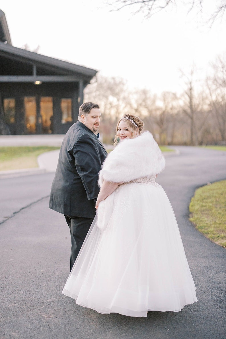 May include: A bride and groom walking down a paved path. The bride is wearing a white wedding dress with a fur shawl and a tiara. The groom is wearing a black suit. They are walking away from the camera and smiling.