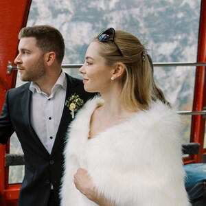 May include: A couple poses for a photo in a cable car. The woman is wearing a white faux fur shawl and the man is wearing a black suit. The couple is looking out the window at the snowy mountain scenery.