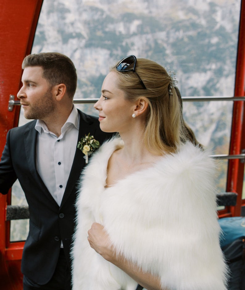 May include: A couple poses for a photo in a cable car. The woman is wearing a white faux fur shawl and the man is wearing a black suit. The couple is looking out the window at the snowy mountain scenery.