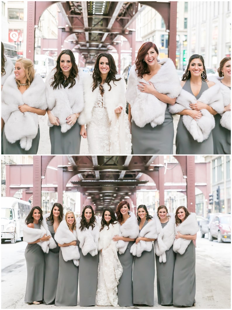 May include: A group of bridesmaids wearing gray dresses and white fur wraps stand in front of a red brick building. The bride is in the center wearing a white lace dress.