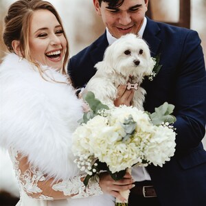 May include: A bride and groom pose for a wedding photo with their small white dog. The bride is wearing a white dress with a fur shawl and holding a bouquet of white flowers. The groom is wearing a dark blue suit and holding the dog.