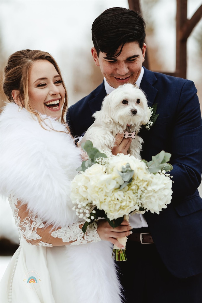 May include: A bride and groom pose for a wedding photo with their small white dog. The bride is wearing a white dress with a fur shawl and holding a bouquet of white flowers. The groom is wearing a dark blue suit and holding the dog.