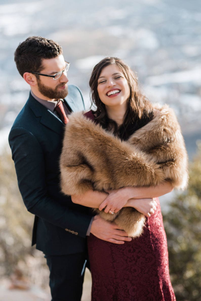 May include: A couple poses for a photo, the woman is wearing a burgundy dress with a brown faux fur shawl, the man is wearing a dark blue suit and a red tie.