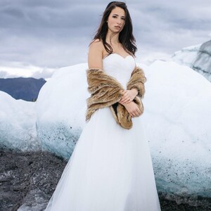 May include: A bride wearing a white wedding dress with a brown fur shawl standing on a glacier.