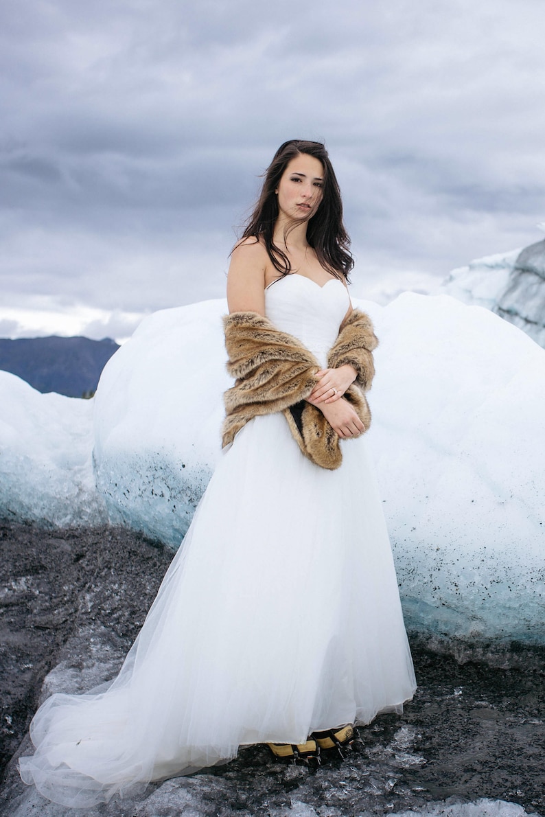 May include: A bride wearing a white wedding dress with a brown fur shawl standing on a glacier.