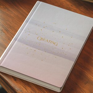 May include: A hardcover journal with a soft color palette of pink, lavender, and white. The word "CREATING" is printed in gold on the cover, with gold dots scattered across the design. The journal is resting on a wooden surface.