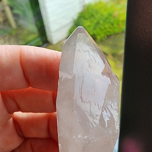 Può includere: Una punta di cristallo di quarzo trasparente con una texture naturale e ruvida. Il cristallo è traslucido e ha una leggera sfumatura rosa.