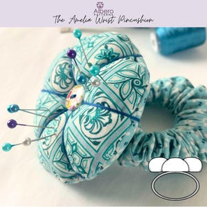 May include: A teal and white patterned wrist pincushion with colourful pins inserted. The pincushion is round and has a button in the centre. The text "The Amelia Wrist Pincushion" is at the top.