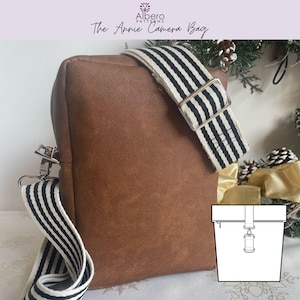 May include: A brown camera bag with a striped strap and silver hardware. The bag has a rectangular shape with a zip closure and a diagram of the bag's design. The bag is labelled "The Annie Camera Bag".