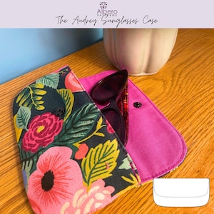 May include: The Audrey Sunglasses Case, open to reveal sunglasses. The case is dark with a floral pattern of pink, yellow, and green flowers. The interior is bright pink. The text "The Audrey Sunglasses Case" is at the top.
