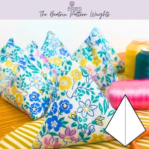 Peut inclure: Des poids de patron triangulaires en tissu avec un motif floral bleu, jaune et rose. Les poids sont disposés sur une surface rayée, avec des bobines de fil et un schéma de patron en arrière-plan. Le texte "The Beatrix Pattern Weights" est en haut.
