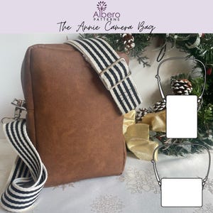 May include: A brown camera bag with a detachable, adjustable shoulder strap featuring black and white horizontal stripes. The bag has a rectangular shape with silver-toned metal hardware. Text at the top reads Albero Patterns The Annie Camera Bag.