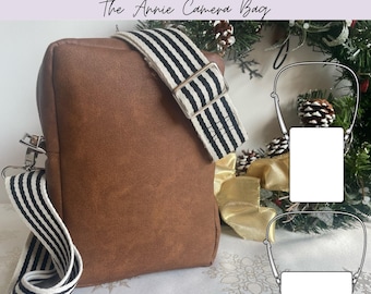 Può includere: Una borsa per fotocamera marrone con tracolla regolabile e rimovibile a righe orizzontali bianche e nere. La borsa ha una forma rettangolare con finiture in metallo argentato. Il testo in alto recita Albero Patterns The Annie Camera Bag.
