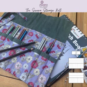 May include: A fabric storage roll featuring a floral pattern in shades of purple, pink, and white, with dark green trim and pockets holding various colored pencils and brushes. The roll includes a tie closure and a diagram showing construction.